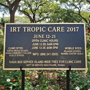 Puede incluir: Un letrero negro y dorado para IRT Tropic Care 2017, con texto que detalla las fechas, horarios y ubicaciones de la clínica. El letrero está rodeado de flores coloridas y vegetación, con un árbol al fondo.