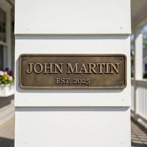Puede incluir: Una placa rectangular de metal en tono bronce con un borde elevado y cuatro tornillos en las esquinas. La placa presenta el texto en relieve JOHN MARTIN en una fuente serif, con el texto EST. 2025 centrado justo debajo en un tamaño menor.