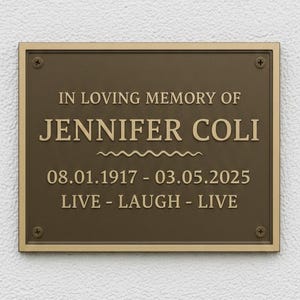 Può includere: Targa rettangolare in bronzo con l'iscrizione "IN LOVING MEMORY OF JENNIFER COLI" e le date "08.01.1917 - 03.05.2025". Le parole "LIVE - LAUGH - LIVE" sono sotto. Montata su un muro bianco strutturato.