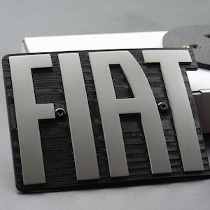 Op de afbeelding: Een rechthoekig Fiat-embleem met zilveren letters op een zwarte, getextureerde achtergrond. Het embleem staat naast een klein, rond, donkergrijs stuk en een witte doos. Het embleem is waarschijnlijk voor een auto.