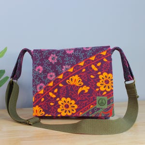 May include: A square crossbody bag with a floral pattern in shades of purple, orange, and yellow. The bag has a green strap and a peace sign patch. A small potted plant sits to the left of the bag.