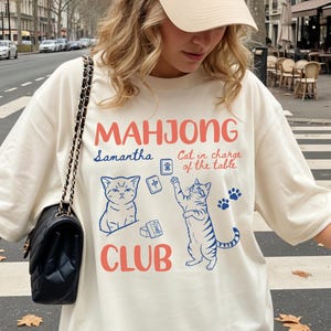May include: An ivory t-shirt featuring the red text MAHJONG CLUB and blue illustrations of two cats, mahjong tiles, and the text Samantha and Cat in charge of the table. The shirt is paired with a beige baseball cap with a small dark logo.