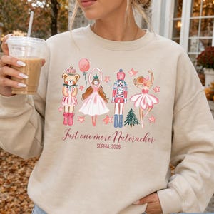 May include: A sand-colored crewneck sweatshirt featuring a graphic print of a teddy bear, a ballerina, a nutcracker, and a mouse dancer with stars and a tree. The text reads Just one more Nutcracker and SOPHIA, 2026 in a script and block font.