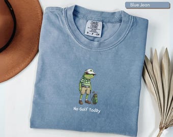 May include: Blue jean colored t-shirt with a graphic of a frog dressed in golf attire, including a hat, striped shirt, and shorts. The text "No Golf Today" is printed below the frog. The shirt has a crew neck and short sleeves.