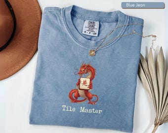 May include: Blue jean colored t-shirt with a red dragon graphic holding a tile. The text "Tile Master" is below the dragon. A gold necklace is draped across the shirt. The shirt has a Comfort Colors tag.