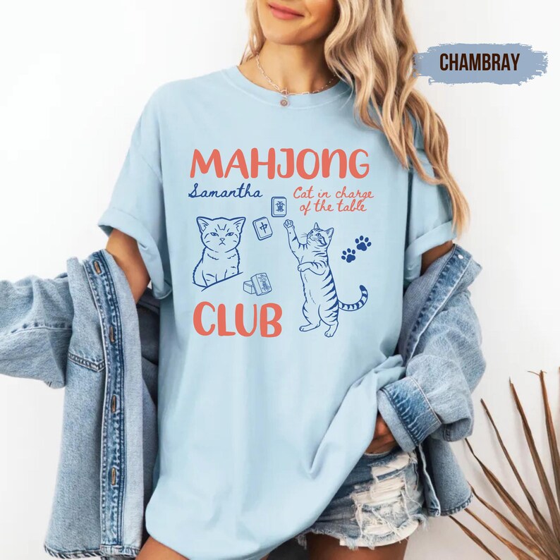 May include: A light blue t-shirt featuring a graphic print with the text "MAHJONG CLUB" in orange, "Samantha" and "Cat in charge of the table" in blue, along with illustrations of two cats and mahjong tiles in blue ink.