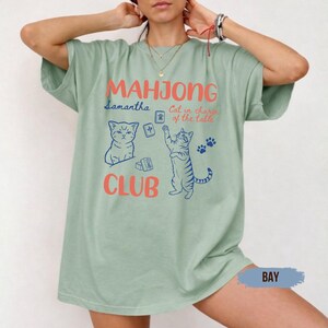 May include: A sage green t-shirt featuring a graphic print with the text "MAHJONG CLUB" in coral, "Samantha" in blue, and "Cat in charge of the table" in coral. The design includes two illustrated cats and several mahjong tiles in blue.