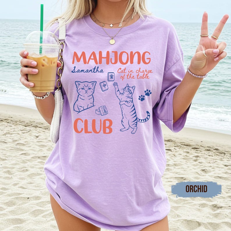 May include: An orchid-coloured t-shirt featuring the text "MAHJONG CLUB" in orange, with blue illustrations of two cats and mahjong tiles. The text "Samantha" and "Cat in charge of the table" are printed in blue script alongside paw print graphics.