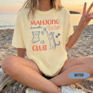 May include: A pale yellow t-shirt featuring the red text "MAHJONG CLUB" and blue illustrations of two cats playing with mahjong tiles. The design includes the blue text "Samantha" and "Cat in charge of the table" with small blue paw prints.