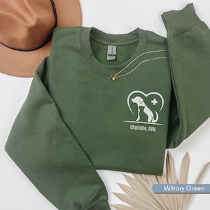 May include: Military green crewneck sweatshirt with a white graphic of a dog and cat inside a heart, with the text "Charlotte, DVM". A gold necklace and a brown hat are also visible.