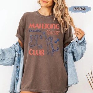 May include: An espresso-coloured t-shirt featuring the text "MAHJONG CLUB" in coral, with blue line art illustrations of two cats, mahjong tiles, and the text "Samantha" and "Cat in charge of the table" printed on the front of the garment.