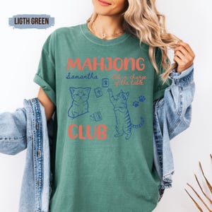 May include: A light green short-sleeve t-shirt featuring the text "MAHJONG CLUB" in orange, with blue illustrations of two cats and mahjong tiles. The shirt also includes the text "Samantha" and "Cat in charge of the table" in blue script.