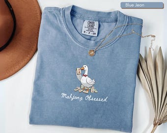May include: Blue jean colored t-shirt with a cartoon duck holding a Mahjong tile. The text "Mahjong Obsessed" is embroidered below the duck. A gold necklace and a brown hat are also in the image.