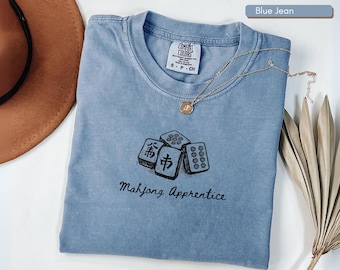 May include: Blue jean colored t-shirt with a graphic of Mahjong tiles and the text "Mahjong Apprentice". A gold necklace with a pendant rests on the shirt. The shirt is made of a soft material.