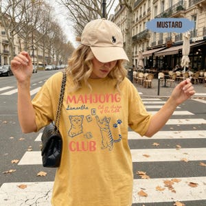 May include: A mustard yellow t-shirt featuring the text "MAHJONG CLUB" in pink, with blue illustrations of two cats, mahjong tiles, and paw prints. The text "Samantha" and "Cat in charge of the table" are printed in blue. A beige baseball cap is worn.