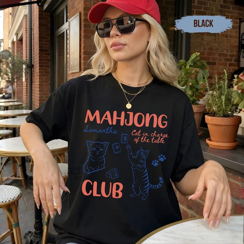 May include: A black t-shirt featuring the text "MAHJONG CLUB" in coral, with blue line art illustrations of two cats, mahjong tiles, and the text "Samantha" and "Cat in charge of the table" in a script font, along with blue paw prints.