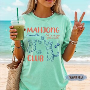 May include: A mint green short-sleeve t-shirt featuring a graphic print with the text "MAHJONG CLUB" in coral, "Samantha" in blue, and "Cat in charge of the table" in coral. The design includes two blue cat illustrations and mahjong tiles.