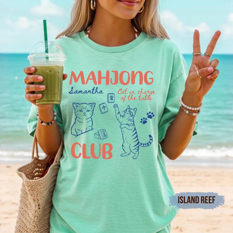 May include: A mint green short-sleeve t-shirt featuring a graphic print with the text "MAHJONG CLUB" in coral, "Samantha" in blue, and "Cat in charge of the table" in coral. The design includes two blue cat illustrations and mahjong tiles.