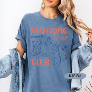 May include: A blue jean coloured t-shirt featuring the orange text MAHJONG CLUB and blue line art of two cats playing with mahjong tiles. The shirt includes the text Samantha and Cat in charge of the table in a blue script font with paw prints.