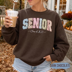 May include: A dark brown long-sleeve sweatshirt featuring the word SENIOR in large, block-style letters. Each letter has a unique pattern, including gingham, floral, and polka dot prints. Below the word, the text Class of 2027 is printed in white.