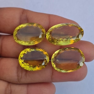 May include: Four oval-cut, faceted gemstones with a yellow-green hue. The gems are displayed against a neutral background, showcasing their clarity and brilliance. The stones are likely for jewelry making.