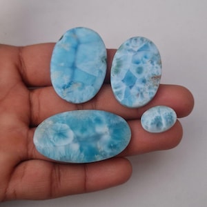 Puede incluir: Cuatro piedras preciosas de larimar ovaladas de diferentes tamaños, que muestran un tono azul claro con patrones blancos y azul oscuro. Las piedras pulidas muestran vetas naturales.