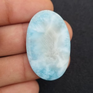 Puede incluir: Una piedra preciosa cabujón de forma ovalada con una superficie lisa y pulida que presenta un patrón jaspeado en colores azul cielo, turquesa y blanco. La piedra muestra un aspecto nublado y translúcido con vetas azul claro y oscuro.