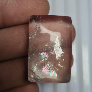 Puede incluir: Una gema rectangular y translúcida con un tono marrón rojizo. La piedra muestra fracturas internas y destellos iridiscentes de colores del arcoíris. La gema se sostiene entre los dedos, mostrando su superficie pulida.