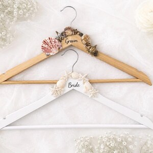 May include: Two decorative wooden wedding hangers. One is natural wood with "Groom" and seashell accents. The other is white with "Bride" and starfish accents. Both have silver metal hooks.