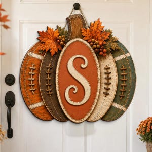 May include: A decorative door hanger shaped like a pumpkin with football-inspired lacing details. The center features an oval plaque with a large cream-colored letter S. It is accented with artificial orange maple leaves and small brown berries.