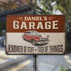 Pode incluir: Uma placa de metal retangular com acabamento desgastado apresentando um carro antigo vermelho ao centro. O texto DANIEL'S GARAGE está no topo e KNOWER OF STUFF FIXER OF THINGS na parte inferior em letras castanho-escuras sobre fundo bicolor.