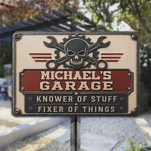 Pode incluir: Uma placa retangular de metal com uma caveira preta centralizada sobre duas chaves inglesas cruzadas e uma engrenagem. O texto diz MICHAEL'S GARAGE, KNOWER OF STUFF, FIXER OF THINGS. A placa tem um fundo creme, uma faixa vermelha e quatro parafusos.