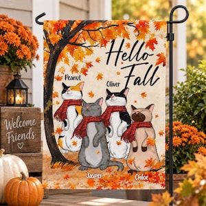 Puede incluir: Una bandera de jardín rectangular con el texto Hello Fall en letra cursiva negra. Cuatro gatos ilustrados con bufandas de cuadros rojos están bajo un árbol de otoño con hojas naranjas. Los gatos llevan los nombres Peanut, Oliver, Jasper y Chloe.