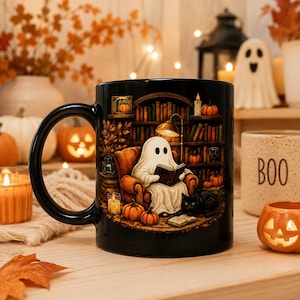 May include: A black ceramic mug featuring a detailed illustration of a white ghost sitting in an orange armchair reading a book, accompanied by a black cat, surrounded by bookshelves, lit candles, and small orange pumpkins in a cozy setting.