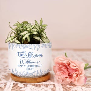 May include: A white ceramic planter with blue floral designs and the text "time bloom to William HAPPY RETIREMENT 2025". The planter holds a small green plant and sits on a wooden coaster. A pink flower rests beside it.