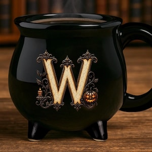 May include: A black ceramic mug shaped like a cauldron with three small feet and a handle. The front features a large, ornate, cream-colored letter W with a cracked texture, surrounded by decorative scrollwork, a small lit candle, and a pumpkin.