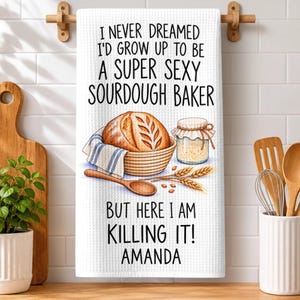 May include: A white waffle-textured kitchen towel featuring a brown sourdough loaf in a basket, a glass jar of starter, and wheat stalks. The text reads: I NEVER DREAMED I'D GROW UP TO BE A SUPER SEXY SOURDOUGH BAKER BUT HERE I AM KILLING IT! AMANDA.