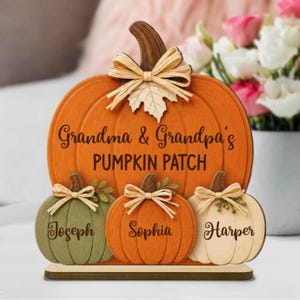 Könnte beinhalten: Ein kürbisförmiges Holzschild mit der Aufschrift Grandma & Grandpa's PUMPKIN PATCH. Darunter befinden sich drei kleinere Kürbisse in Olivgrün, Orange und Creme, die mit den Namen Joseph, Sophia und Harper beschriftet und mit Bastschleifen verziert sind.