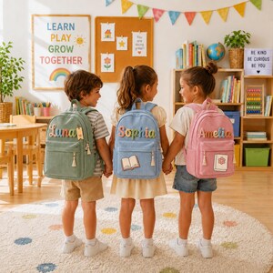 Puede incluir: Tres niños llevan mochilas de pana en verde salvia, azul claro y rosa empolvado. Las mochilas presentan los nombres bordados Luna, Sophia y Amelia, junto con pequeños parches de un libro y un libro decorado con una flor.