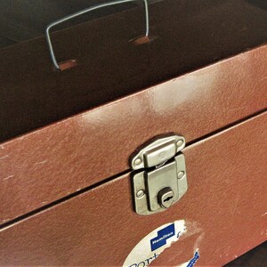 May include: A brown rectangular storage case with a metallic silver handle on top and a silver metal latch closure on the front. The case features a partially torn white circular sticker with the blue text Hamilton and partial blue lettering below.