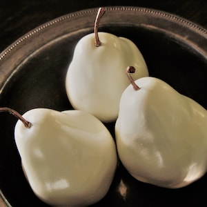 May include: Three white ceramic pears in a silver bowl. The pears are smooth and glossy, with a slight sheen.
