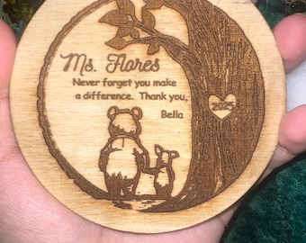 Puede incluir: Adorno navideño redondo de madera con un diseño de Winnie the Pooh y Piglet, un árbol y el texto "Ms. Flores Never forget you make a difference. Thank you, Bella 2025". Tiene un lazo de cuerda.