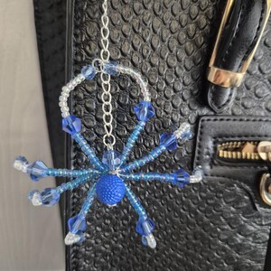 May include: A beaded spider ornament featuring a textured blue central bead, eight legs crafted from light blue and clear glass beads, and a silver-toned metal chain for hanging. The spider is constructed with wire and various faceted blue beads.