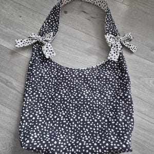 May include: A navy blue tote bag with a white polka dot pattern. The bag has a curved top and two straps with bows. The interior and straps are white with black polka dots. The bag is made of fabric.