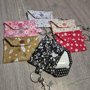 May include: A collection of six small fabric pouches with keychains, each featuring a different pattern including floral, star, and polka dot designs in colours like pink, yellow, black, and white, with snap button closures and metal hardware.