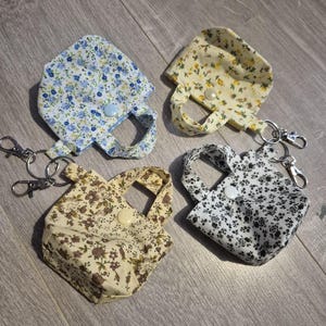 May include: Four small fabric tote bag keyrings. Each bag features a different floral pattern: blue, yellow, tan, and black and white. Each bag has a small handle and a button closure. The keyrings are attached with silver-coloured clasps.