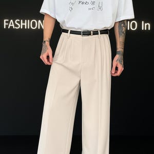 May include: Beige pleated trousers with a black belt and white sneakers. The top is a white t-shirt with a small graphic and the text "Feb.18 202*" in black. The word "FASHION" is visible on the left.