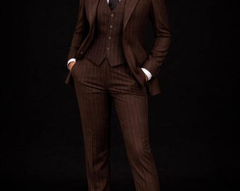 Pode incluir: Um fato castanho risca de giz, composto por blazer, colete e calças, usado com uma camisa branca e uma gravata preta. A imagem também mostra sapatos de salto alto pretos. O logótipo "FB FASHION BLOOM -STUDIO-" está no canto superior direito.