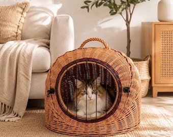 May include: A round, light brown woven wicker pet carrier with a top handle and a circular front opening secured by a black metal grate. A long-haired tabby cat with white and brown fur is sitting inside the enclosure, looking forward.