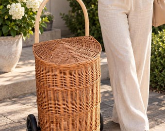 May include: A tall, cylindrical wicker shopping cart with a natural light brown finish, featuring a curved wooden handle, a hinged lid, and two black wheels at the base for mobility. The woven basket structure is detailed with horizontal bands.